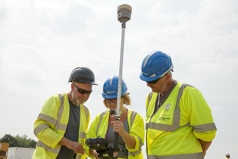 Three people in high vis PPE are looking at the screen of a total station. The total station is a long white pole with a round section on top.