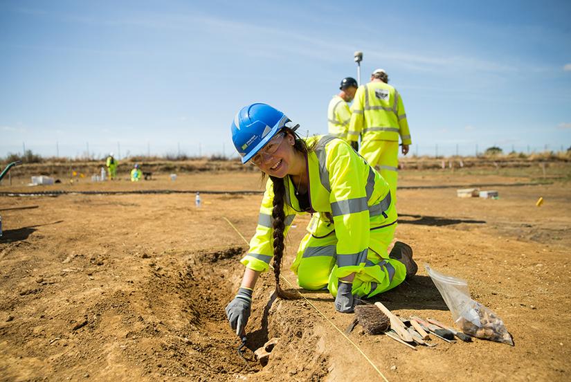 A young woman in high vis PPE and blue hard hat is excavating in an archaeological trench