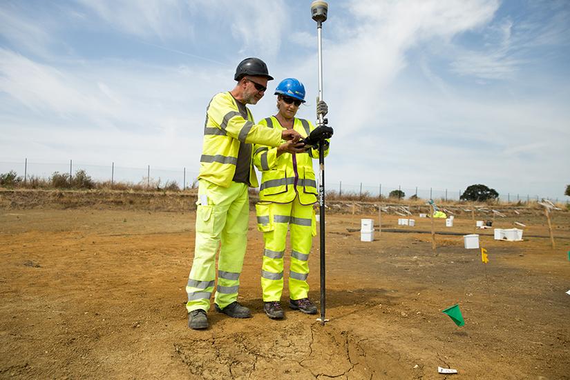 Two people in high vis PPE looking at the screen of a total station. The total station is a long white pole with a round piece on top. They are standing in the middle of a large open archaeological excavation area