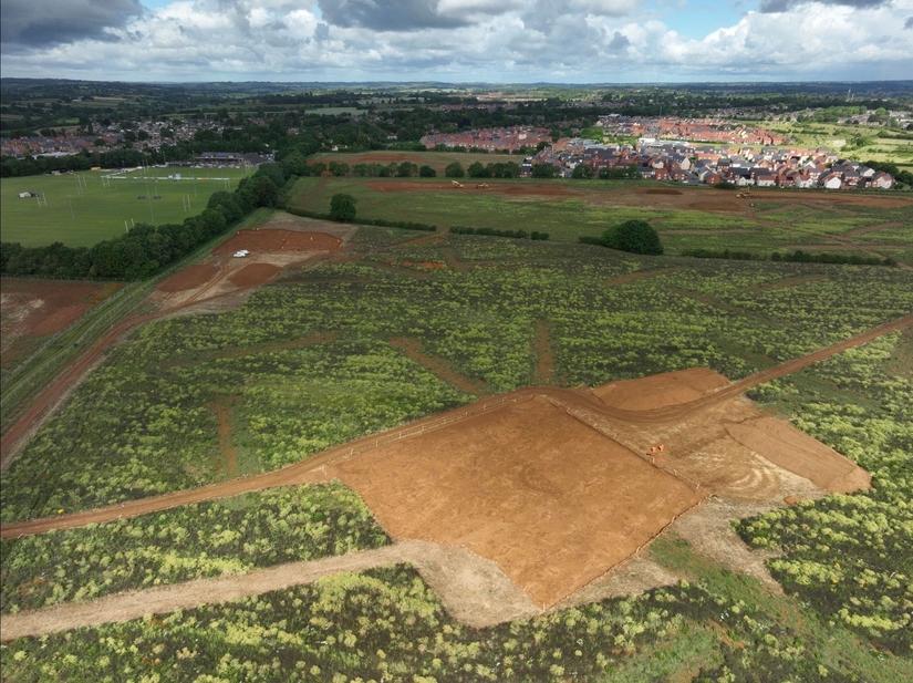 A drone photograph showing an excavated area in the middle of a green field. In the centre of the excavated area is a faint circle