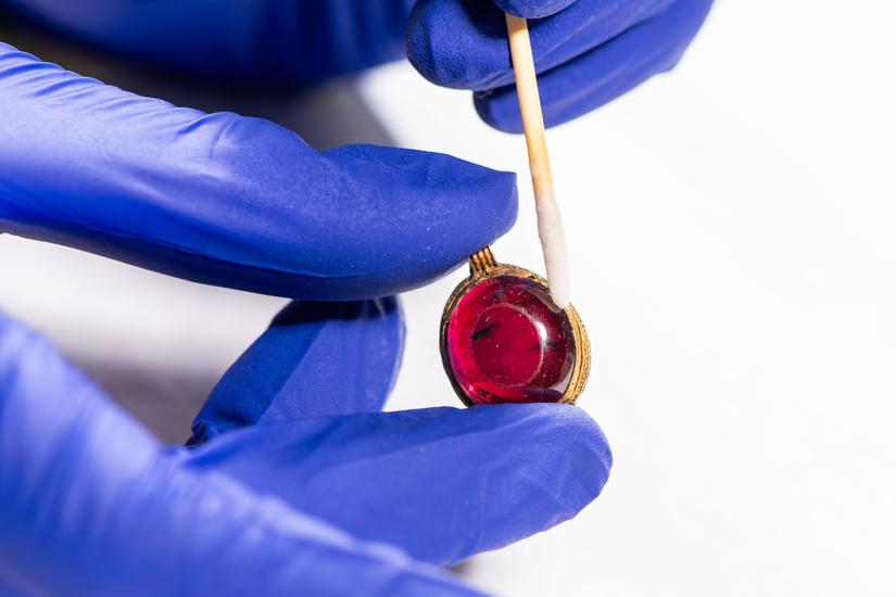 Photo of conservator cleaning a red gemstone pendant, wearing gloves and using a cotton bud