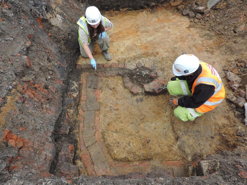 Two archaeologists excavating a brick feature with a small pit filled with rubbish including clay pipes