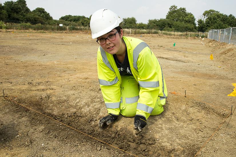 Photograph of a a young woman wearing high vis PPE and a white hard hat using a trowel to excavate in a trench