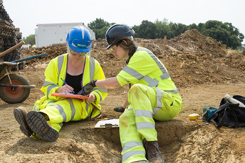 Two people sitting together. One is drawing and the other is pointing at the drawing. Both are dressed in high vis PPE and hard hats