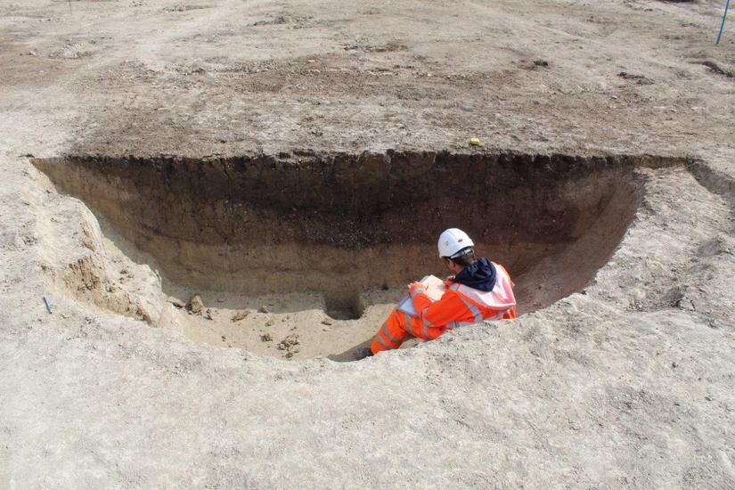 Photo of circular pit, half of which has been excavated and has an archaeologist sat comfortably within