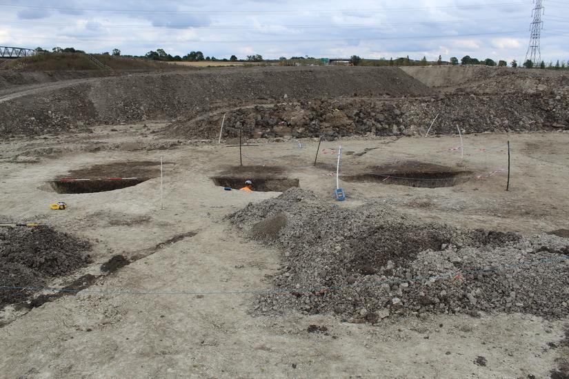 Photo of three pits in a row, you can just see the top of an archaeologist's helmet in one