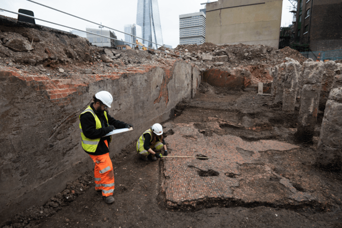 Two archaeologists record a piece of Roman mosaic flooring. They are standing in a large trench.