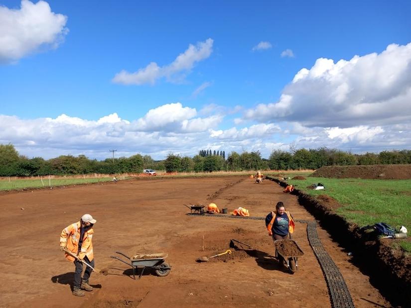 Two archaeologists excavating in an open area in a field on a sunny day. There are more archaeologists excavating in the distance