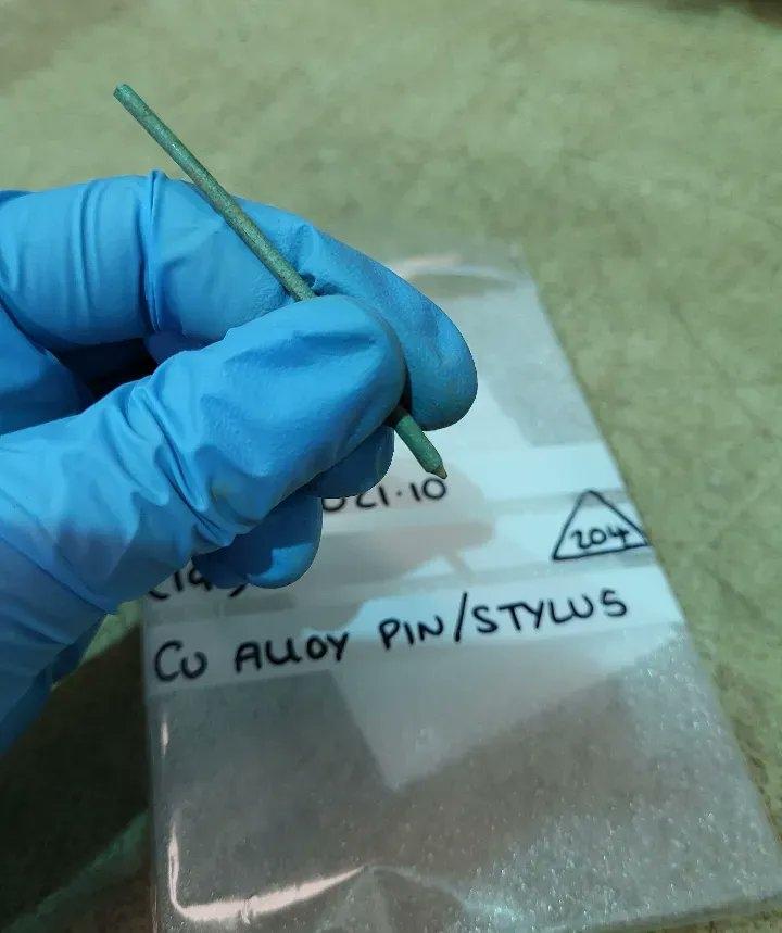 A small pencil-like Roman stylus being held by an archaeologist wearing a blue plastic glove.