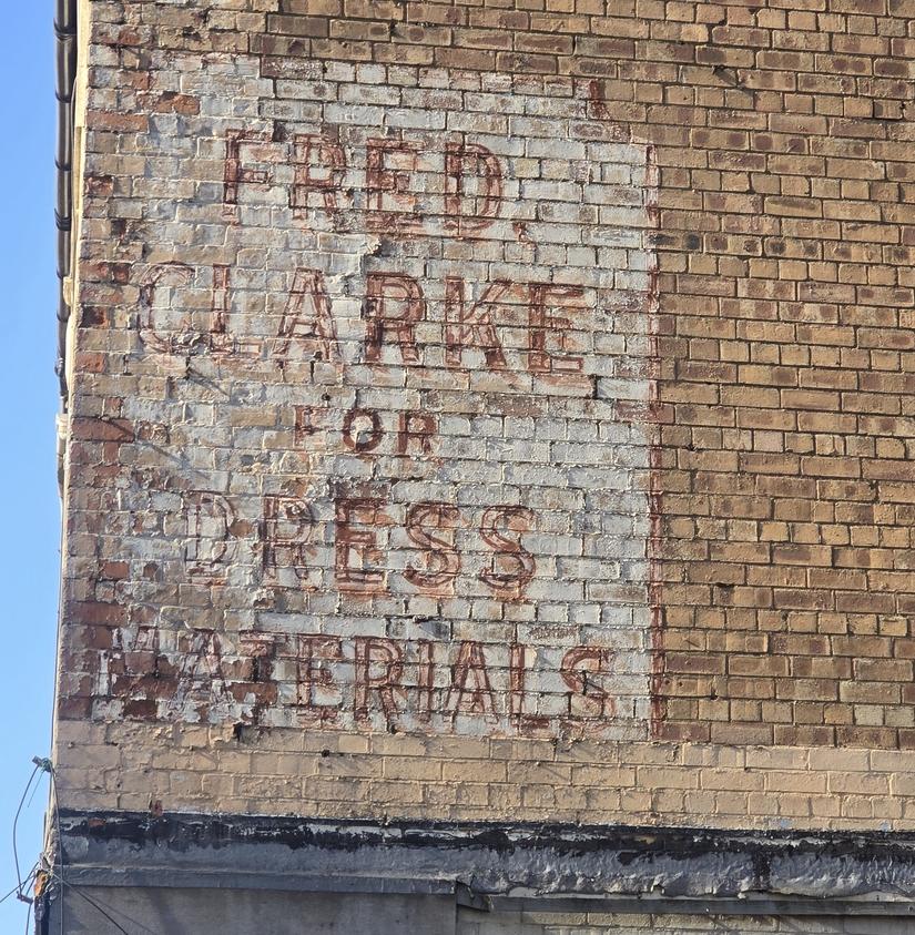 A faded painted sign on a building reads "Fred Clarke for dress materials"