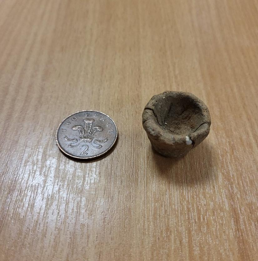 A tiny earth coloured pot on a table. A 2 pence piece is next to it for scale.