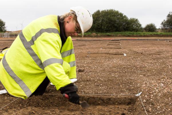 A woman in high vis and a white hard had using a trowel to excavate a square test pit