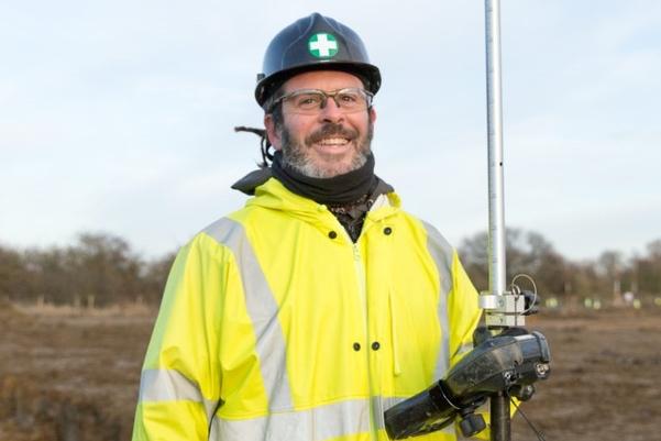 A man in high vis and black hard hat holding a piece of survey equipment