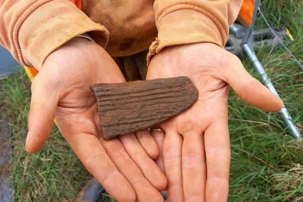 two hands holding a brown piece of pottery decorated with lines around the edges