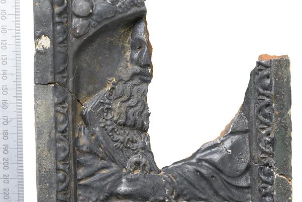 A dark ceramic tile decorated with the image of a man with a large bushy beard. The top right corner of the tile is missing.